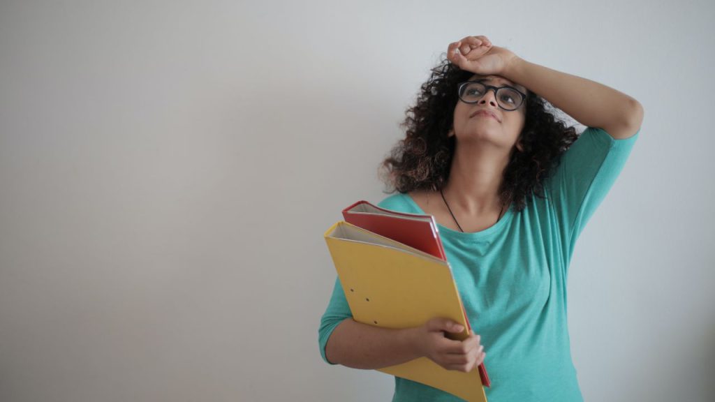 A young Black woman carrying folders of paperwork and looking overwhelmed against a gray background