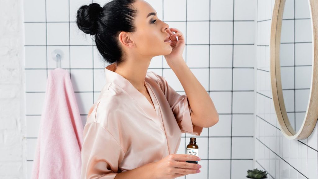 woman in a pink robe applying retinol filler to face in front of mirror