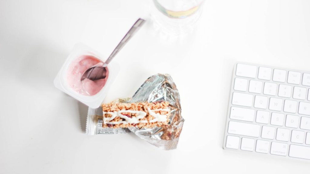 a half-eaten plastic yogurt cup and unwrapped granola bar sit next to a white keyboard against a white background.